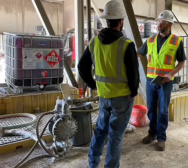 Two men stand in the production area of a composite pipe  manufacturer. In the background is a Sandpiper AODD pump on the ground and several large totes containing a resin solution.